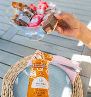 Person holding a caramel snack bar with a bowl of chilled bars on a table outdoors.