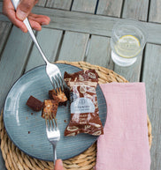 Person holding a fork over a plate with dessert and a chocolate bar, on a wooden table.